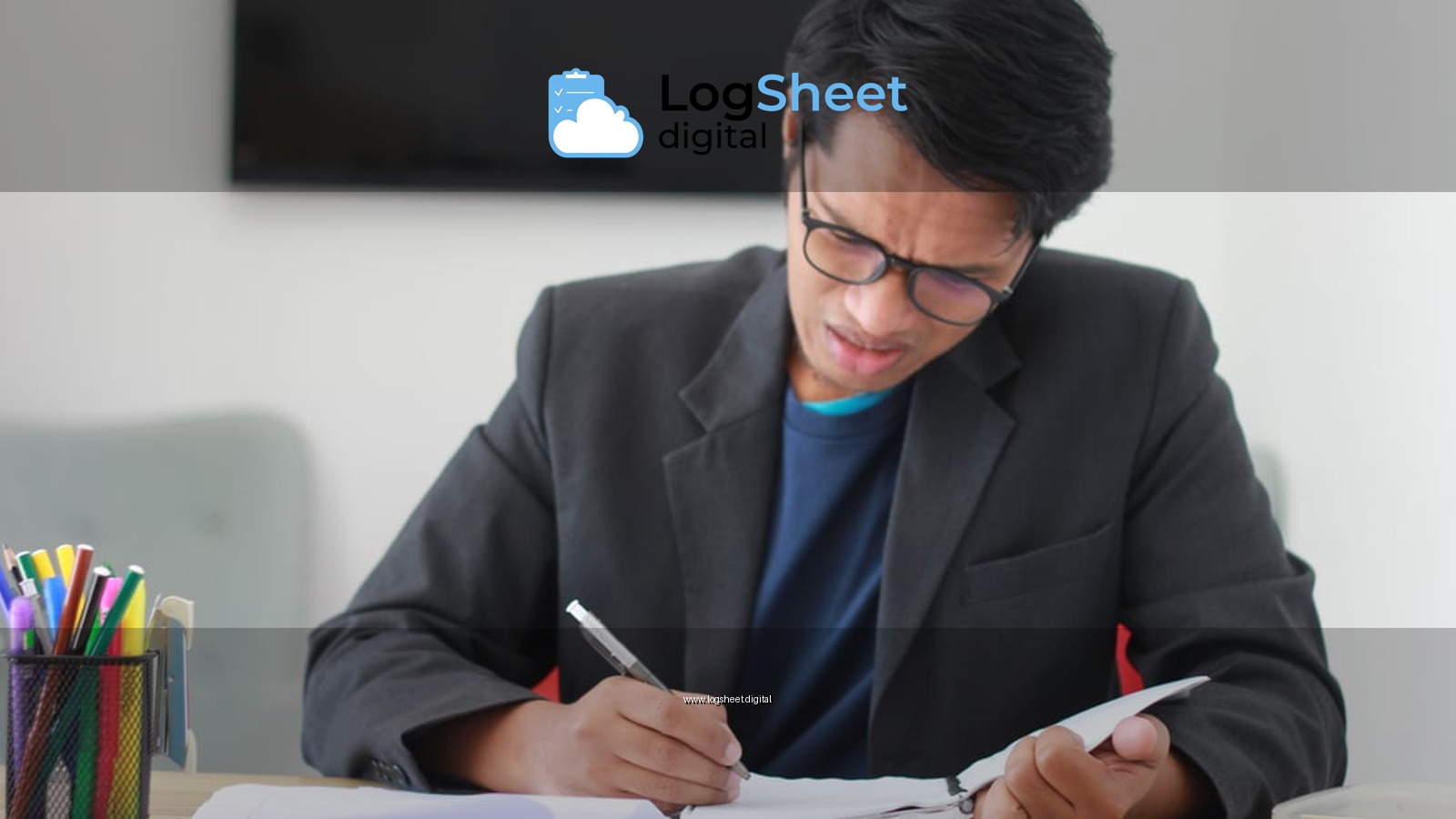 Adult male in a black suit takes notes at a cluttered office desk, looking stressed.