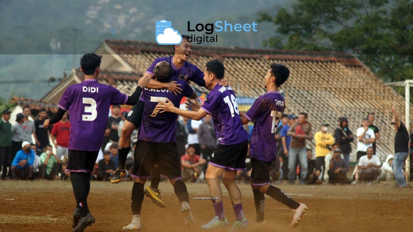 Football players celebrating a goal in a dusty outdoor match with spectators.